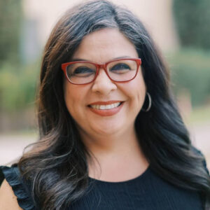 Rose Lucero, wearing dark top and red glasses, smiles with a school blurred in the background.